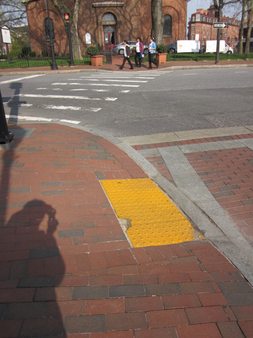 Church Circle: Three photos show a brick crosswalk at a signalized intersection.  The sidewalks are all made of red brick, and at the curb the bricks are laid to form a slope about 4 feet wide down to the street.  Where it meets the street is a detectable warning (DW) about 2 feet deep and 4 feet wide.  The bright yellow surface of the DW has peeled back or eroded at the top of one of the ramps revealing a few inches of white cement.